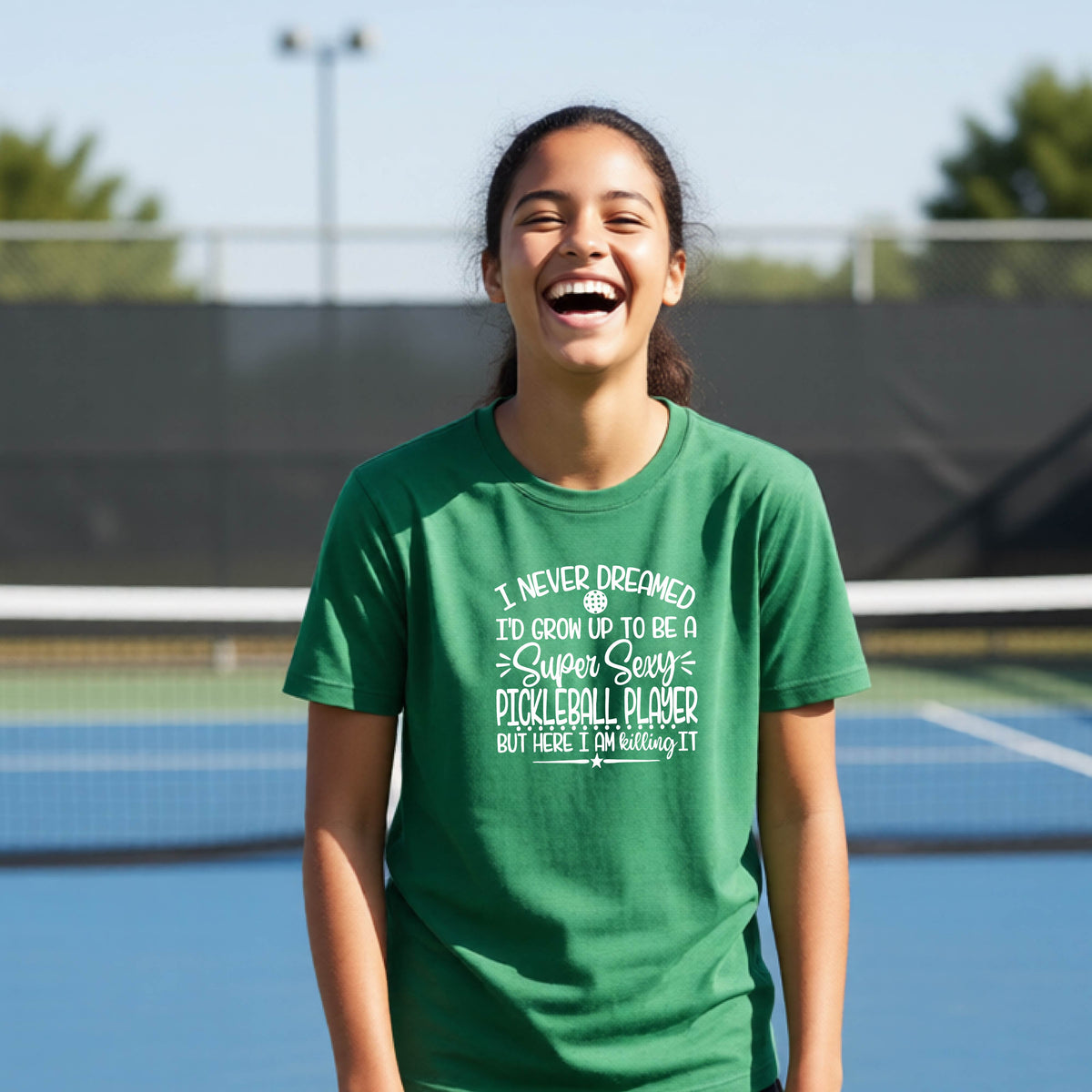A young person on a pickleball court wearing a green long-sleeve t-shirt with a white graphic of the text:  ‘I Never Dreamed I’d Grow Up To Be A Super Sexy Pickleball Player But Here I Am Killing It!’, standing on a pickleball court.