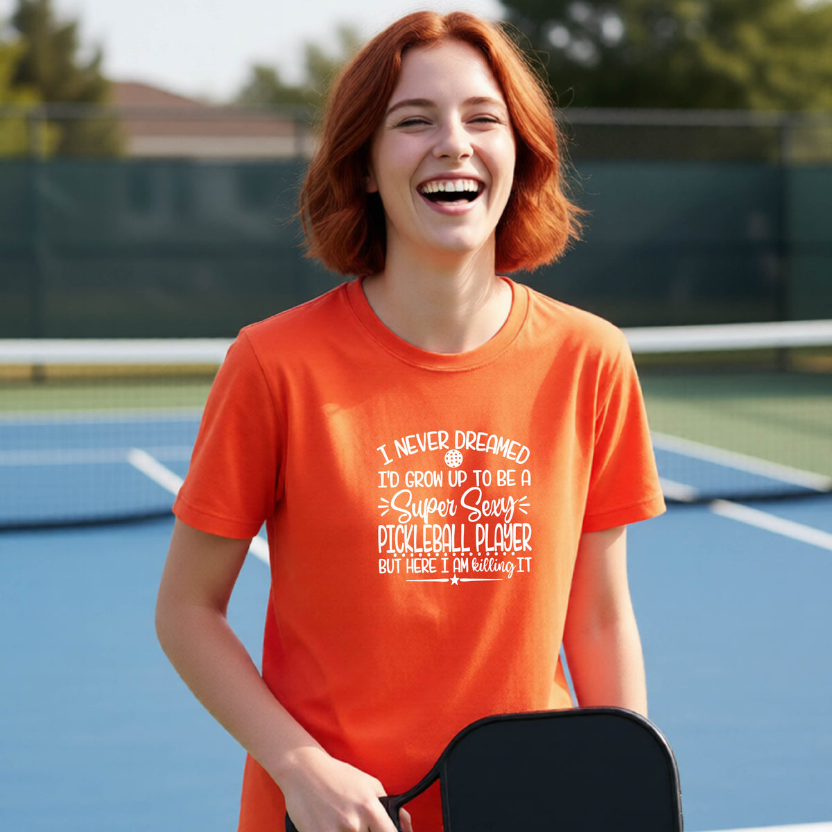 A young person on a pickleball court wearing an orange long-sleeve t-shirt with a white graphic of the text:  ‘I Never Dreamed I’d Grow Up To Be A Super Sexy Pickleball Player But Here I Am Killing It!’, standing on a pickleball court.