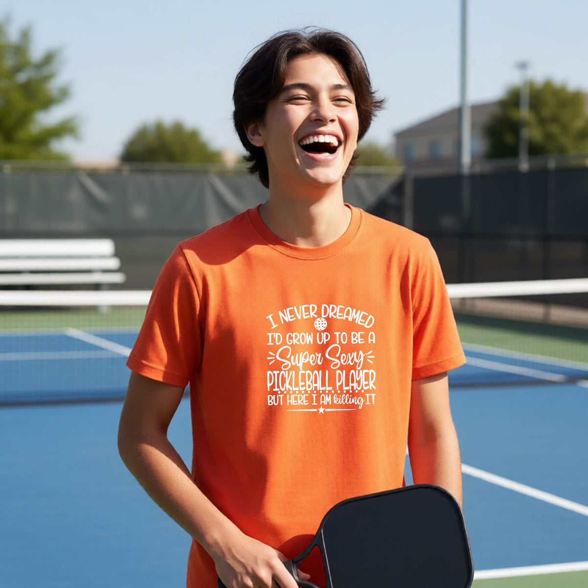 A young person on a pickleball court wearing an orange long-sleeve t-shirt with a white graphic of the text:  ‘I Never Dreamed I’d Grow Up To Be A Super Sexy Pickleball Player But Here I Am Killing It!’, standing on a pickleball court.