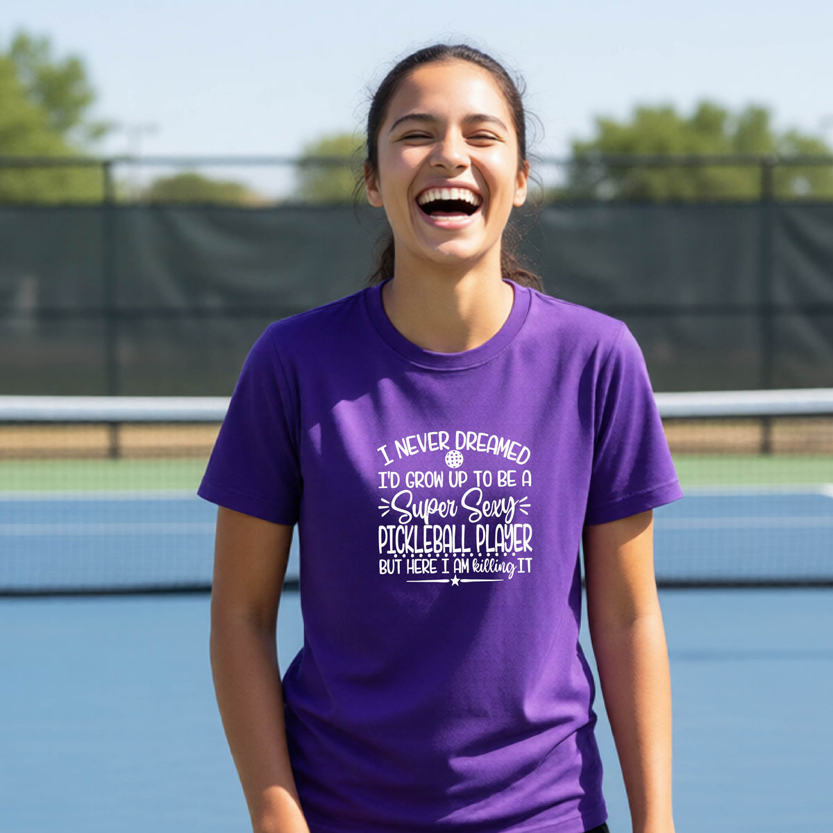 A young person on a pickleball court wearing a purple long-sleeve t-shirt with a white graphic of the text:  ‘I Never Dreamed I’d Grow Up To Be A Super Sexy Pickleball Player But Here I Am Killing It!’, standing on a pickleball court.