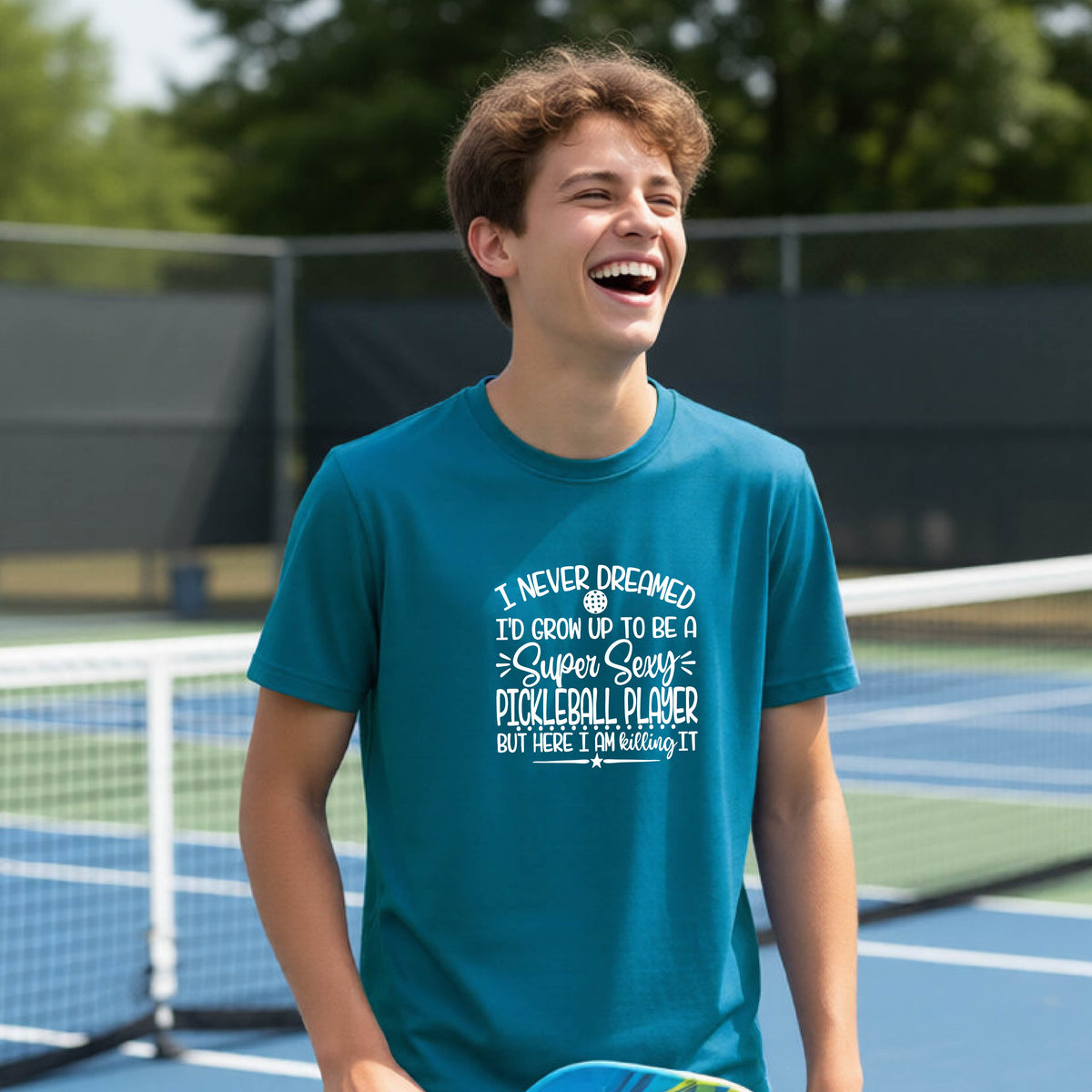 A young person on a pickleball court wearing a teal long-sleeve t-shirt with a white graphic of the text:  ‘I Never Dreamed I’d Grow Up To Be A Super Sexy Pickleball Player But Here I Am Killing It!’, standing on a pickleball court.