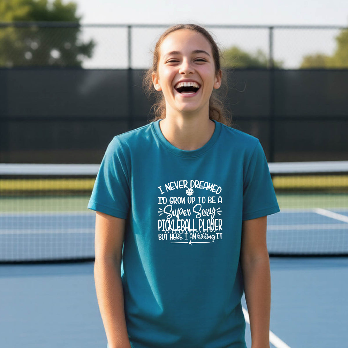 A young person on a pickleball court wearing a teal long-sleeve t-shirt with a white graphic of the text:  ‘I Never Dreamed I’d Grow Up To Be A Super Sexy Pickleball Player But Here I Am Killing It!’, standing on a pickleball court.