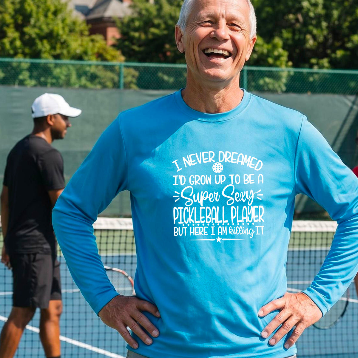 A man on a pickleball court wearing a bright blue long-sleeve t-shirt with a white graphic of the text:  ‘I Never Dreamed I’d Grow Up To Be A Super Sexy Pickleball Player But Here I Am Killing It!’, standing on a pickleball court.