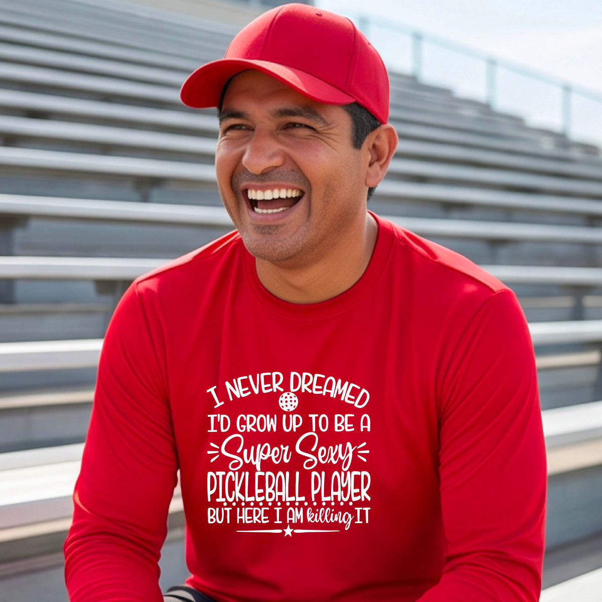 A man on a pickleball court wearing a red long-sleeve t-shirt with a white graphic of the text:  ‘I Never Dreamed I’d Grow Up To Be A Super Sexy Pickleball Player But Here I Am Killing It!’, standing on a pickleball court.