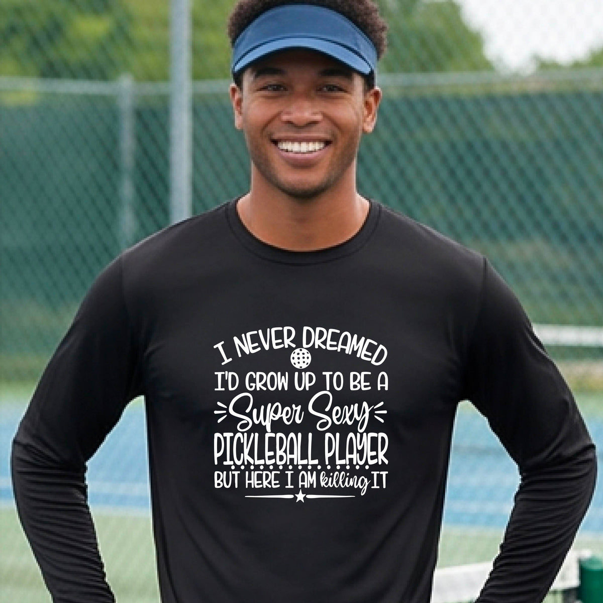 A man on a pickleball court wearing a black long-sleeve t-shirt with a white graphic of the text:  ‘I Never Dreamed I’d Grow Up To Be A Super Sexy Pickleball Player But Here I Am Killing It!’, standing on a pickleball court.