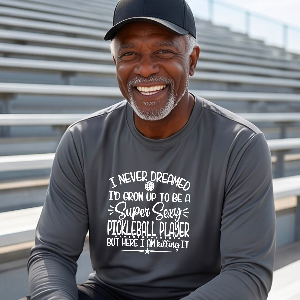 A man on a pickleball court wearing a dark gray long-sleeve t-shirt with a white graphic of the text:  ‘I Never Dreamed I’d Grow Up To Be A Super Sexy Pickleball Player But Here I Am Killing It!’, standing on a pickleball court.