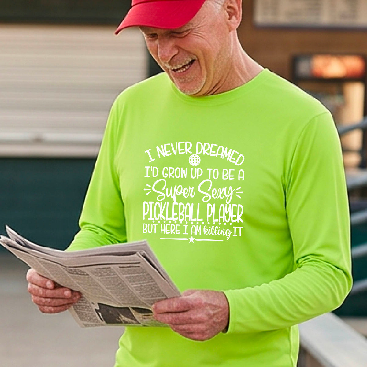 A man on a pickleball court wearing a lime green long-sleeve t-shirt with a white graphic of the text:  ‘I Never Dreamed I’d Grow Up To Be A Super Sexy Pickleball Player But Here I Am Killing It!’, standing on a pickleball court.