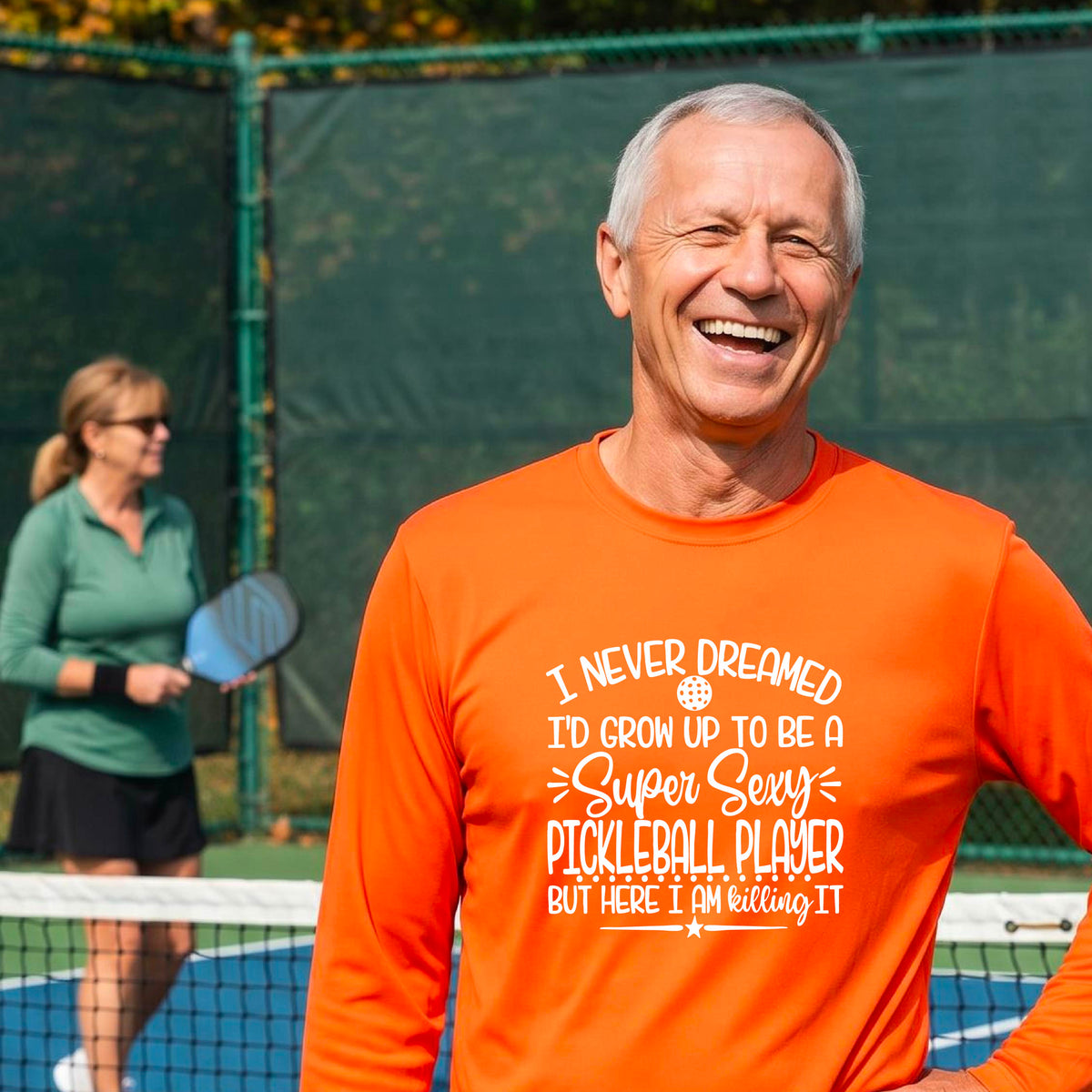 A man on a pickleball court wearing an orange long-sleeve t-shirt with a white graphic of the text:  ‘I Never Dreamed I’d Grow Up To Be A Super Sexy Pickleball Player But Here I Am Killing It!’, standing on a pickleball court.