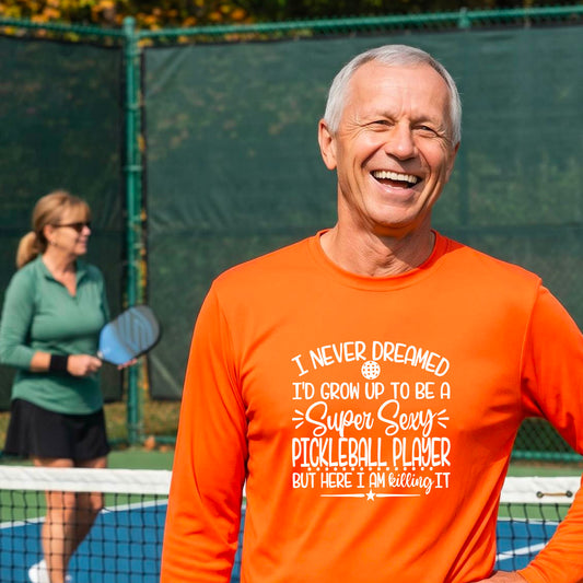 A man on a pickleball court wearing an orange long-sleeve t-shirt with a white graphic of the text:  ‘I Never Dreamed I’d Grow Up To Be A Super Sexy Pickleball Player But Here I Am Killing It!’, standing on a pickleball court.