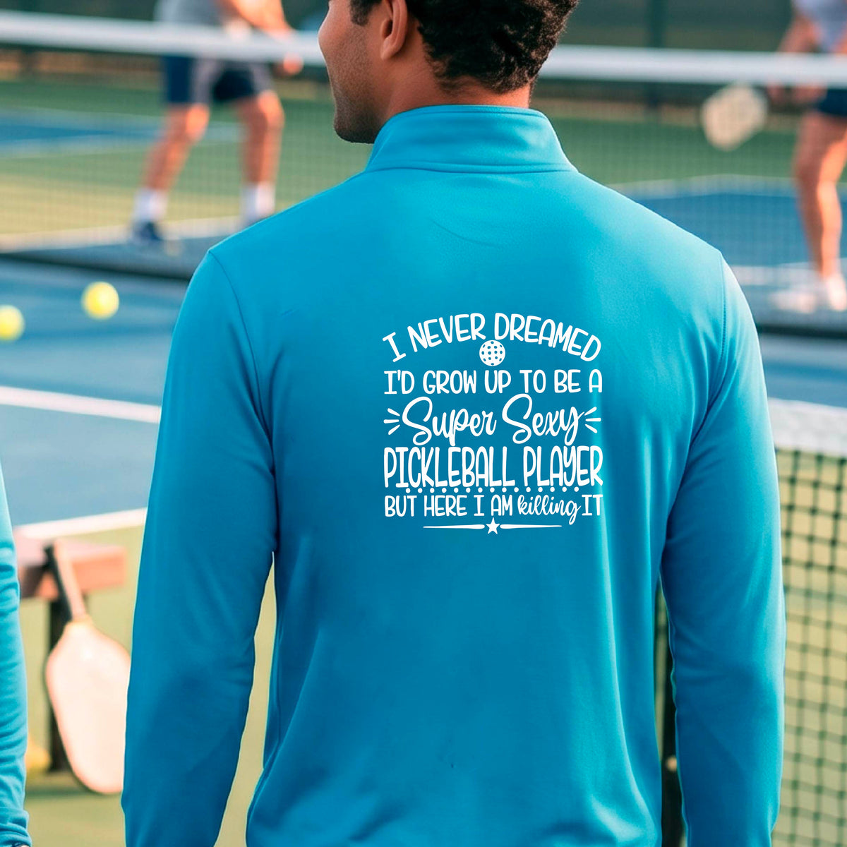 A man on a pickleball court , facing backwards, wearing a bright blue quarter-zip shirt with a white graphic of the text:  ‘I Never Dreamed I’d Grow Up To Be A Super Sexy Pickleball Player But Here I Am Killing It!’, standing on a pickleball court.