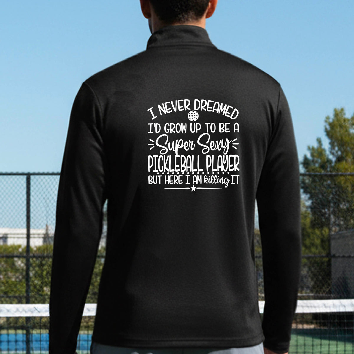 A man on a pickleball court , facing backwards, wearing a black quarter-zip shirt with a white graphic of the text:  ‘I Never Dreamed I’d Grow Up To Be A Super Sexy Pickleball Player But Here I Am Killing It!’, standing on a pickleball court.
