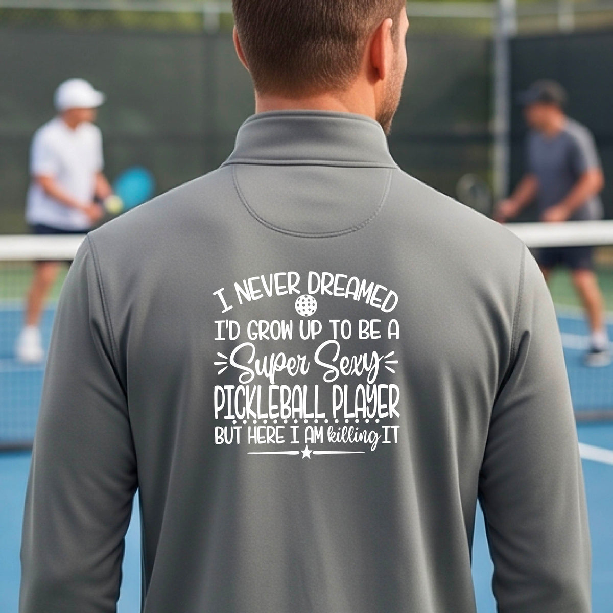 A man on a pickleball court , facing backwards, wearing a gray quarter-zip shirt with a white graphic of the text:  ‘I Never Dreamed I’d Grow Up To Be A Super Sexy Pickleball Player But Here I Am Killing It!’, standing on a pickleball court.