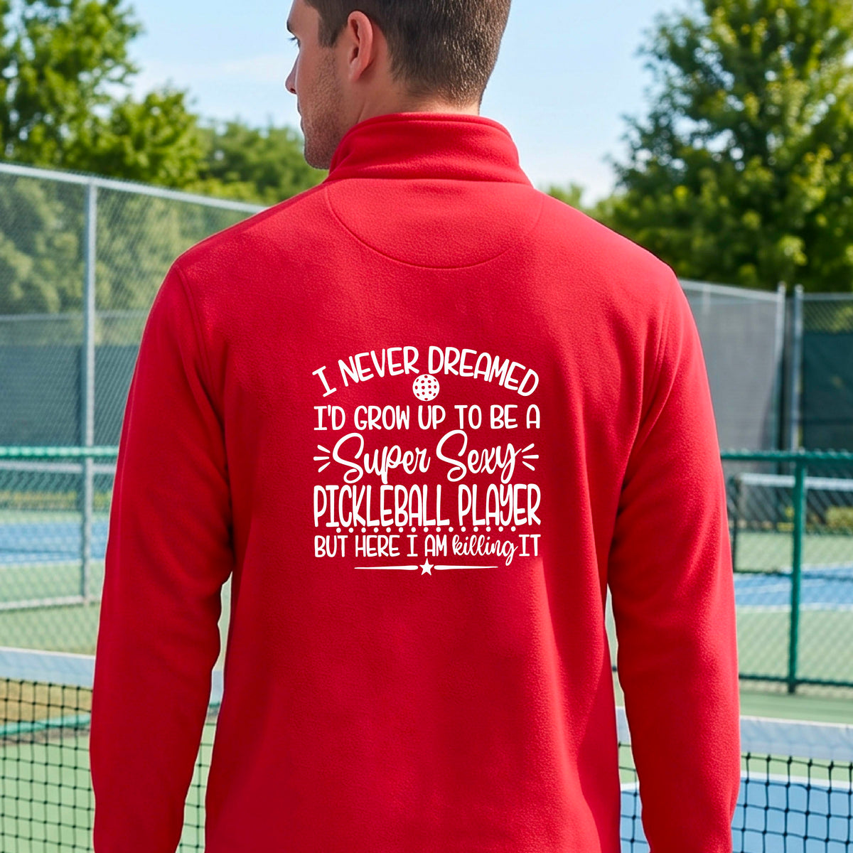 A man on a pickleball court , facing backwards, wearing a red quarter-zip shirt with a white graphic of the text:  ‘I Never Dreamed I’d Grow Up To Be A Super Sexy Pickleball Player But Here I Am Killing It!’, standing on a pickleball court.