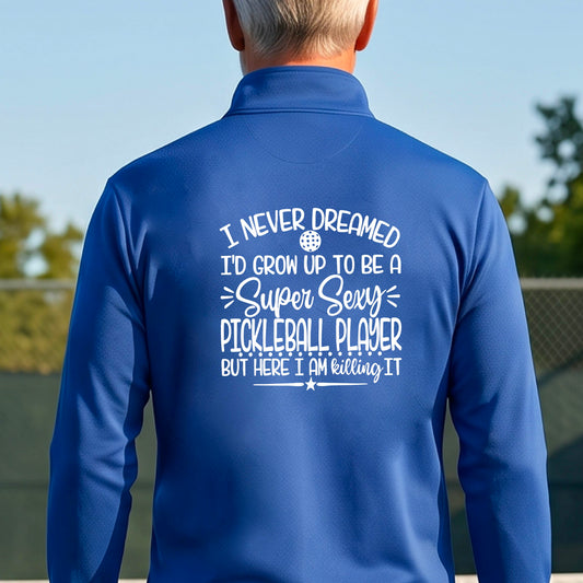 A man on a pickleball court , facing backwards, wearing a black quarter-zip shirt with a white graphic of the text:  ‘I Never Dreamed I’d Grow Up To Be A Super Sexy Pickleball Player But Here I Am Killing It!’, standing on a pickleball court.

