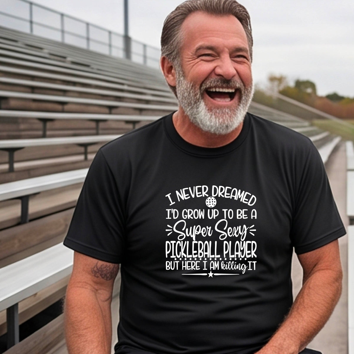 A man on a pickleball court wearing a black short-sleeve t-shirt with a white graphic of the text:  ‘I Never Dreamed I’d Grow Up To Be A Super Sexy Pickleball Player But Here I Am Killing It!’, standing on a pickleball court.