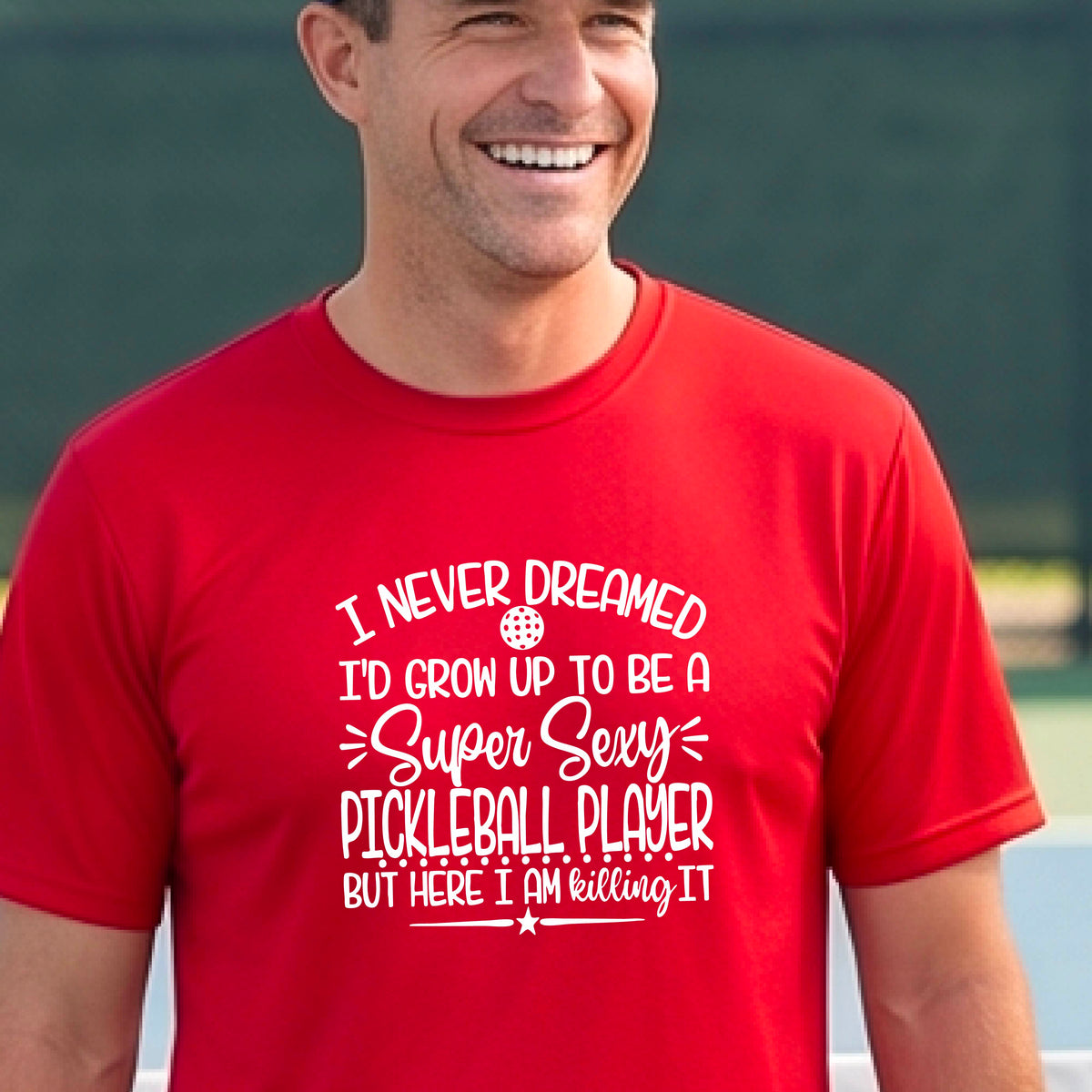 A man on a pickleball court wearing a red short-sleeve t-shirt with a white graphic of the text:  ‘I Never Dreamed I’d Grow Up To Be A Super Sexy Pickleball Player But Here I Am Killing It!’, standing on a pickleball court.