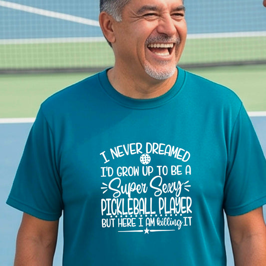 A man on a pickleball court wearing a teal short-sleeve t-shirt with a white graphic of the text:  ‘I Never Dreamed I’d Grow Up To Be A Super Sexy Pickleball Player But Here I Am Killing It!’, standing on a pickleball court.