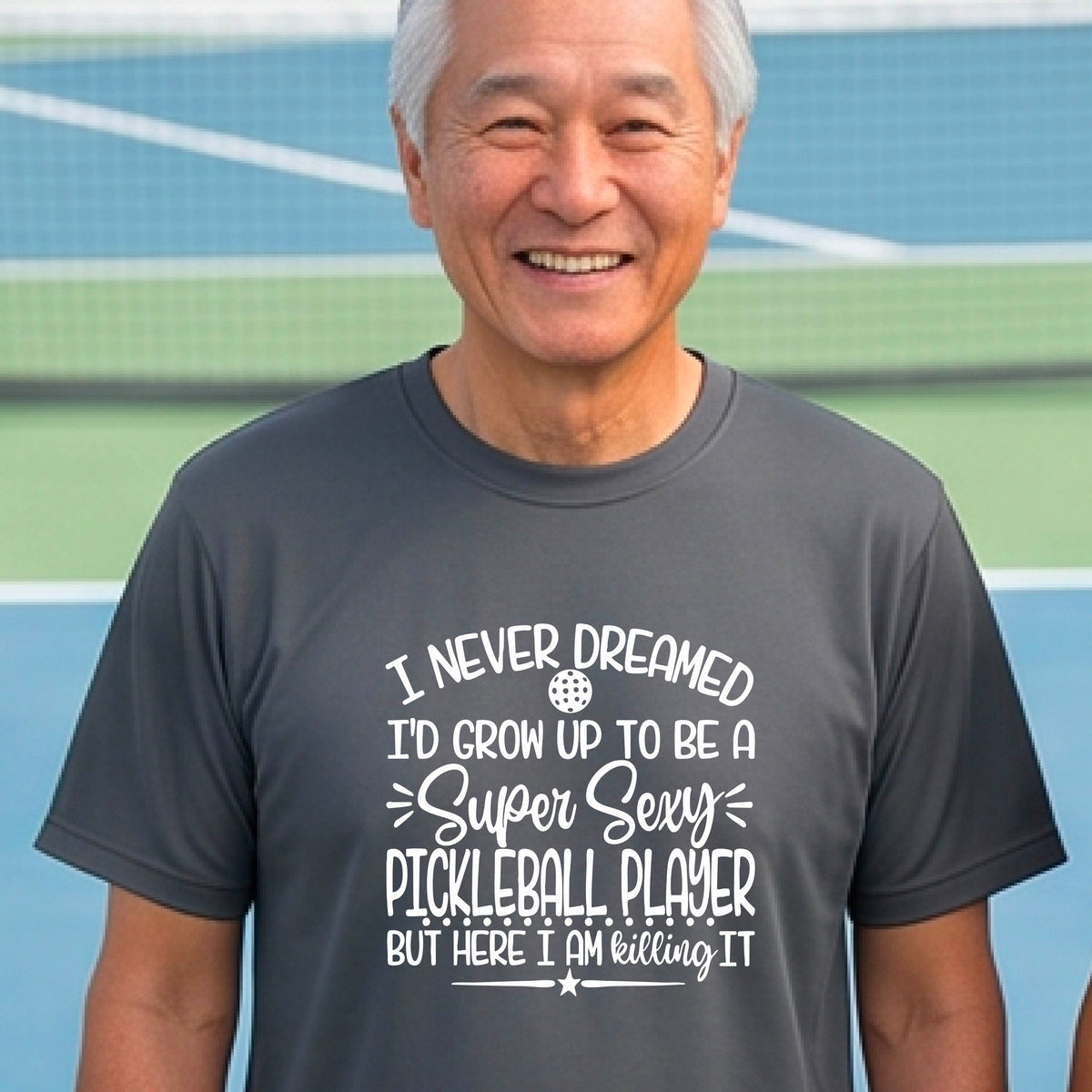 A man on a pickleball court wearing a dark gray short-sleeve t-shirt with a white graphic of the text:  ‘I Never Dreamed I’d Grow Up To Be A Super Sexy Pickleball Player But Here I Am Killing It!’, standing on a pickleball court.