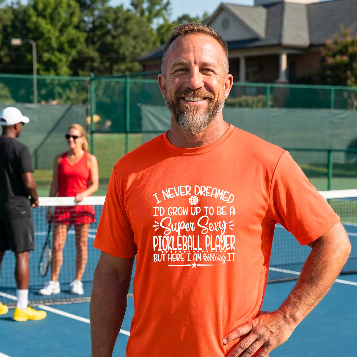 A man on a pickleball court wearing an orange short-sleeve t-shirt with a white graphic of the text:  ‘I Never Dreamed I’d Grow Up To Be A Super Sexy Pickleball Player But Here I Am Killing It!’, standing on a pickleball court.