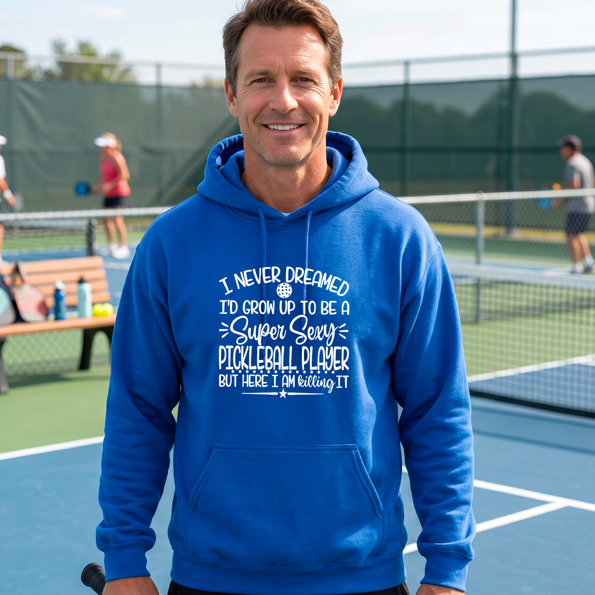 A man on a pickleball court wearing a royal blue unisex hoodie pullover with a white graphic of the text:  ‘I Never Dreamed I’d Grow Up To Be A Super Sexy Pickleball Player But Here I Am Killing It!’, standing on a pickleball court.
