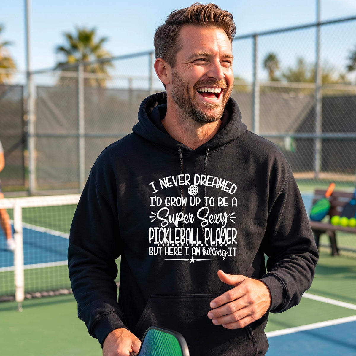 A man on a pickleball court wearing a black unisex hoodie pullover with a white graphic of the text:  ‘I Never Dreamed I’d Grow Up To Be A Super Sexy Pickleball Player But Here I Am Killing It!’, standing on a pickleball court.