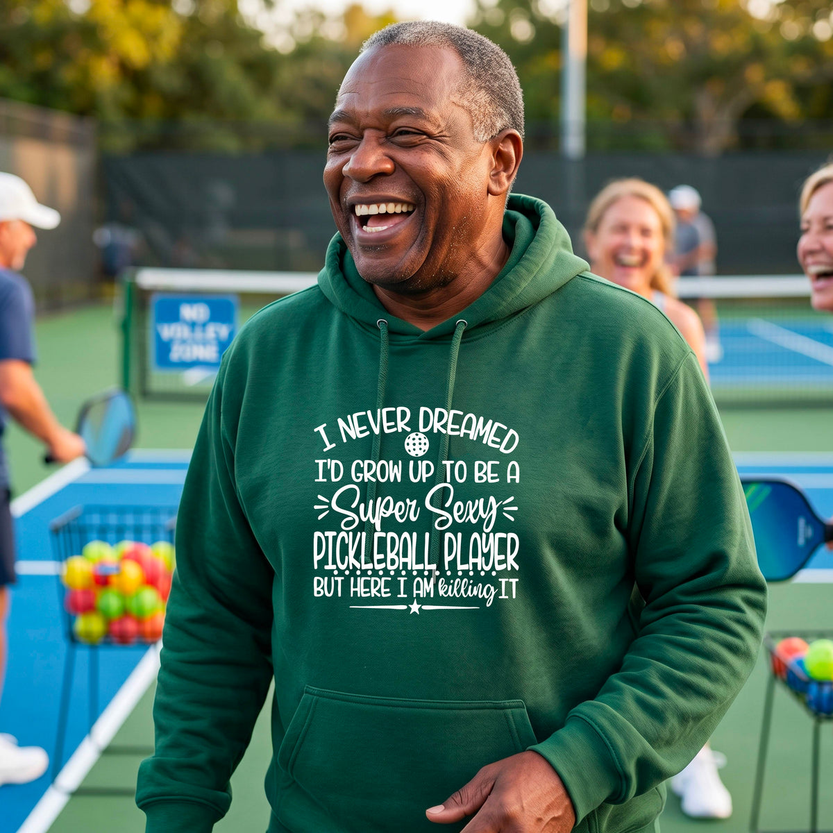 A man on a pickleball court wearing a green unisex hoodie pullover with a white graphic of the text:  ‘I Never Dreamed I’d Grow Up To Be A Super Sexy Pickleball Player But Here I Am Killing It!’, standing on a pickleball court.