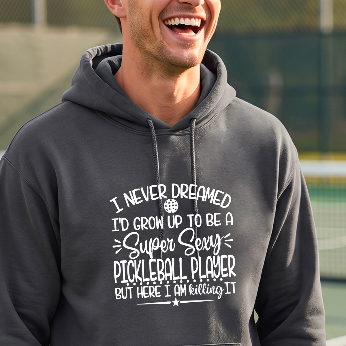 A man on a pickleball court wearing a dark gray unisex hoodie pullover with a white graphic of the text:  ‘I Never Dreamed I’d Grow Up To Be A Super Sexy Pickleball Player But Here I Am Killing It!’, standing on a pickleball court.