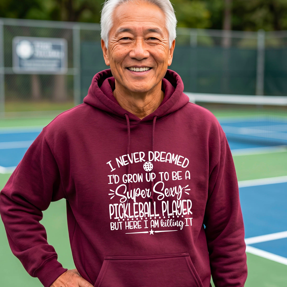 A man on a pickleball court wearing a maroon unisex hoodie pullover with a white graphic of the text:  ‘I Never Dreamed I’d Grow Up To Be A Super Sexy Pickleball Player But Here I Am Killing It!’, standing on a pickleball court.