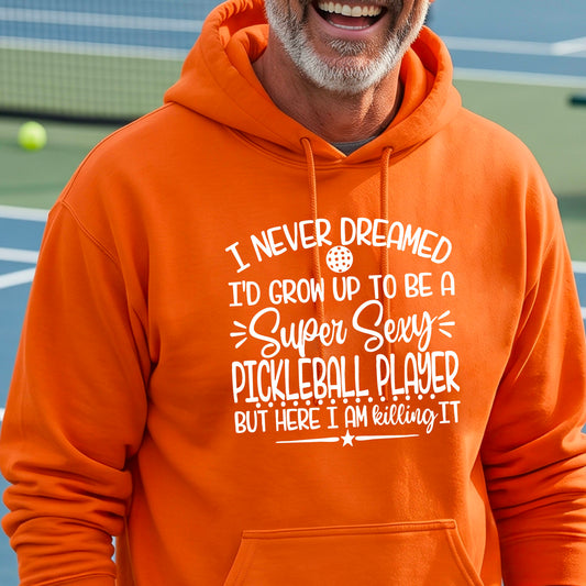 A man on a pickleball court wearing an orange unisex hoodie pullover with a white graphic of the text:  ‘I Never Dreamed I’d Grow Up To Be A Super Sexy Pickleball Player But Here I Am Killing It!’, standing on a pickleball court.
