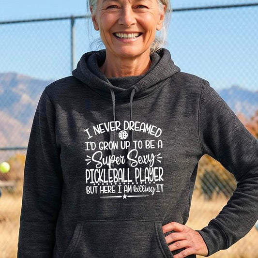 A woman on a pickleball court wearing a black hoodie pullover with a white graphic of the text:  ‘I Never Dreamed I’d Grow Up To Be A Super Sexy Pickleball Player But Here I Am Killing It!’, standing on a pickleball court.