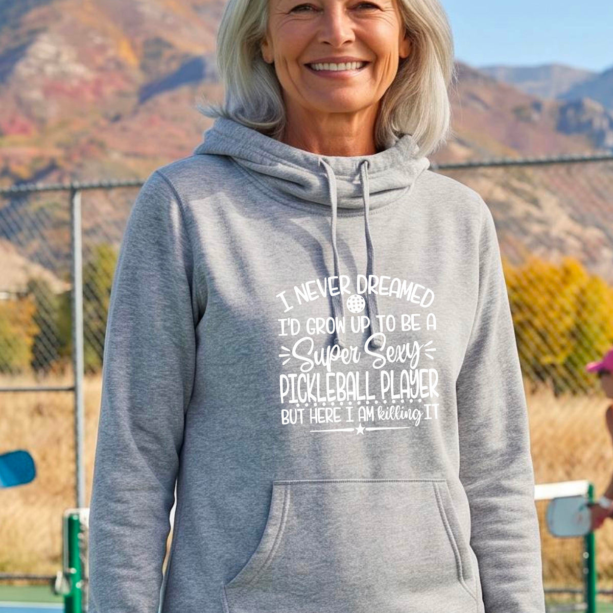 A woman on a pickleball court wearing a light gray hoodie pullover with a white graphic of the text:  ‘I Never Dreamed I’d Grow Up To Be A Super Sexy Pickleball Player But Here I Am Killing It!’, standing on a pickleball court.