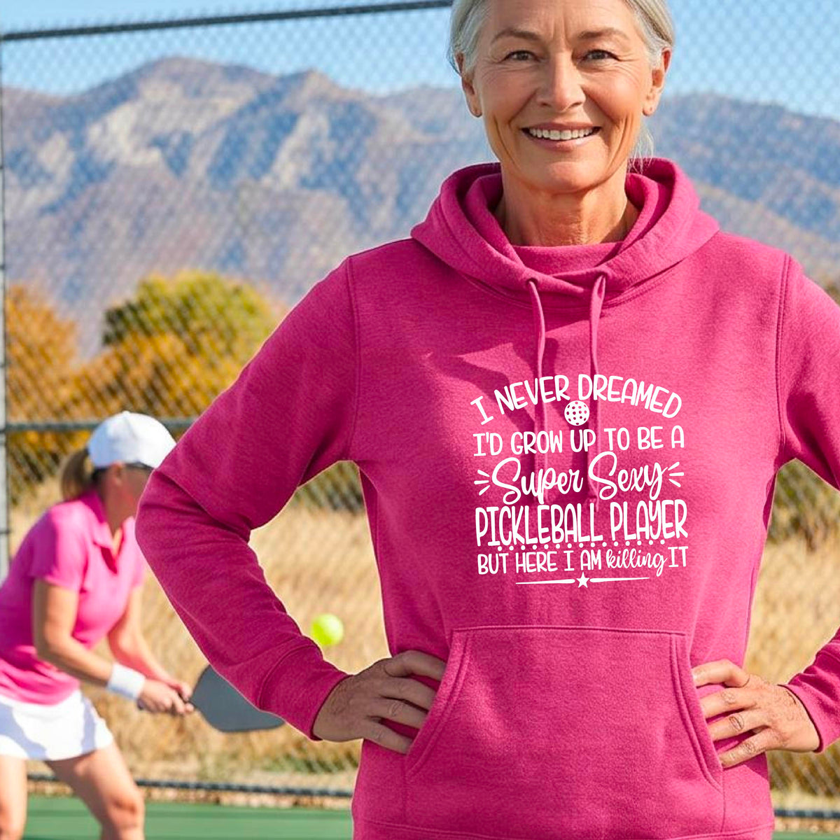 A woman on a pickleball court wearing a pink hoodie pullover with a white graphic of the text:  ‘I Never Dreamed I’d Grow Up To Be A Super Sexy Pickleball Player But Here I Am Killing It!’, standing on a pickleball court.