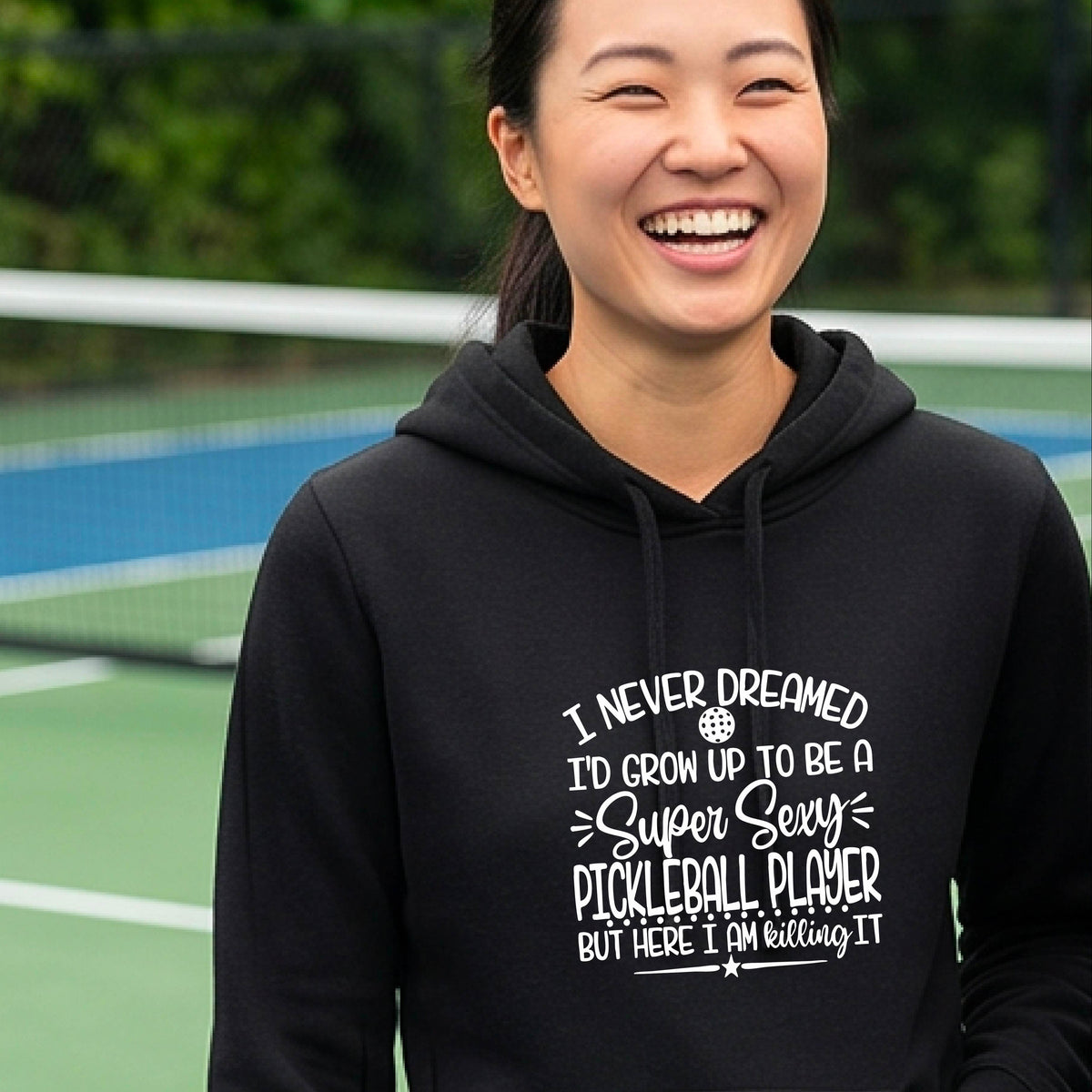 A woman on a pickleball court wearing a black hoodie pullover with a white graphic of the text:  ‘I Never Dreamed I’d Grow Up To Be A Super Sexy Pickleball Player But Here I Am Killing It!’, standing on a pickleball court.