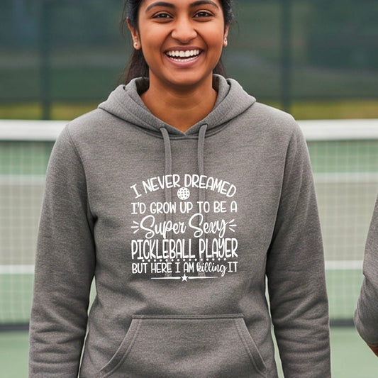 A woman on a pickleball court wearing a black hoodie pullover with a white graphic of the text:  ‘I Never Dreamed I’d Grow Up To Be A Super Sexy Pickleball Player But Here I Am Killing It!’, standing on a pickleball court.

