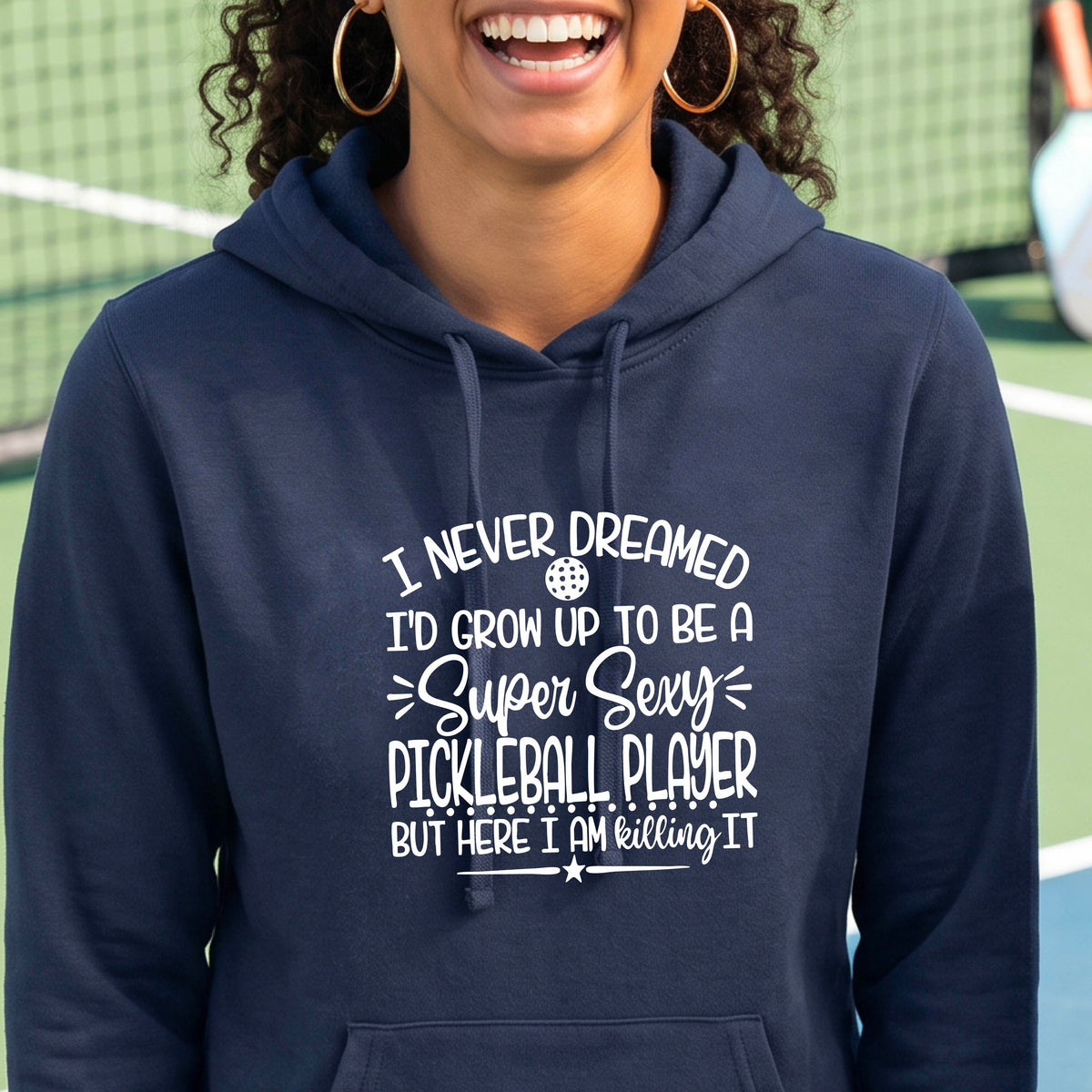A woman on a pickleball court wearing a navy blue hoodie pullover with a white graphic of the text:  ‘I Never Dreamed I’d Grow Up To Be A Super Sexy Pickleball Player But Here I Am Killing It!’, standing on a pickleball court.