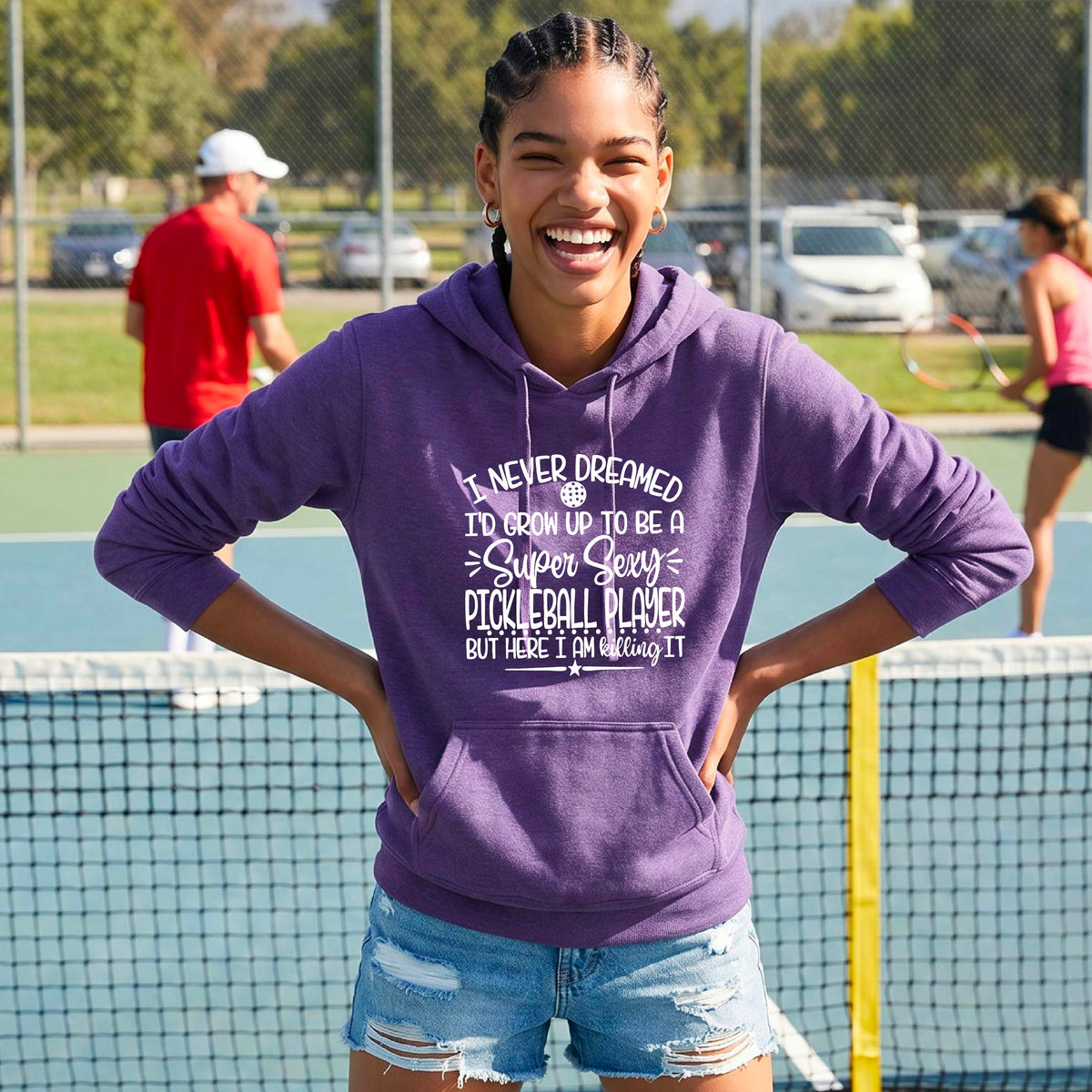 A woman on a pickleball court wearing a purple hoodie pullover with a white graphic of the text:  ‘I Never Dreamed I’d Grow Up To Be A Super Sexy Pickleball Player But Here I Am Killing It!’, standing on a pickleball court.