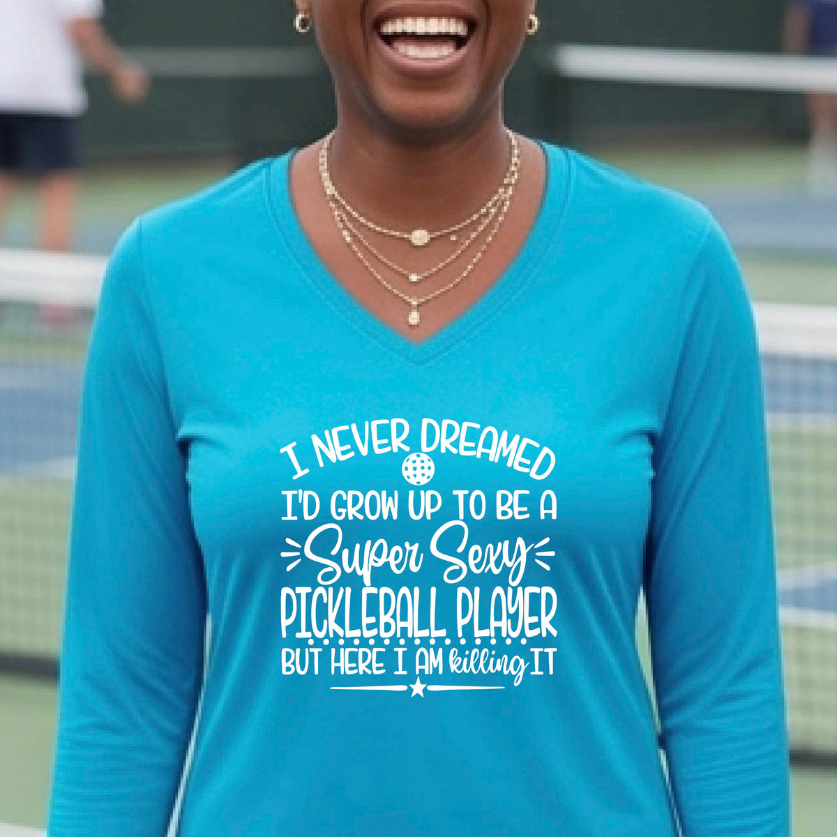 A woman on a pickleball court wearing a bright blue long-sleeve t-shirt with a white graphic of the text:  ‘I Never Dreamed I’d Grow Up To Be A Super Sexy Pickleball Player But Here I Am Killing It!’, standing on a pickleball court.