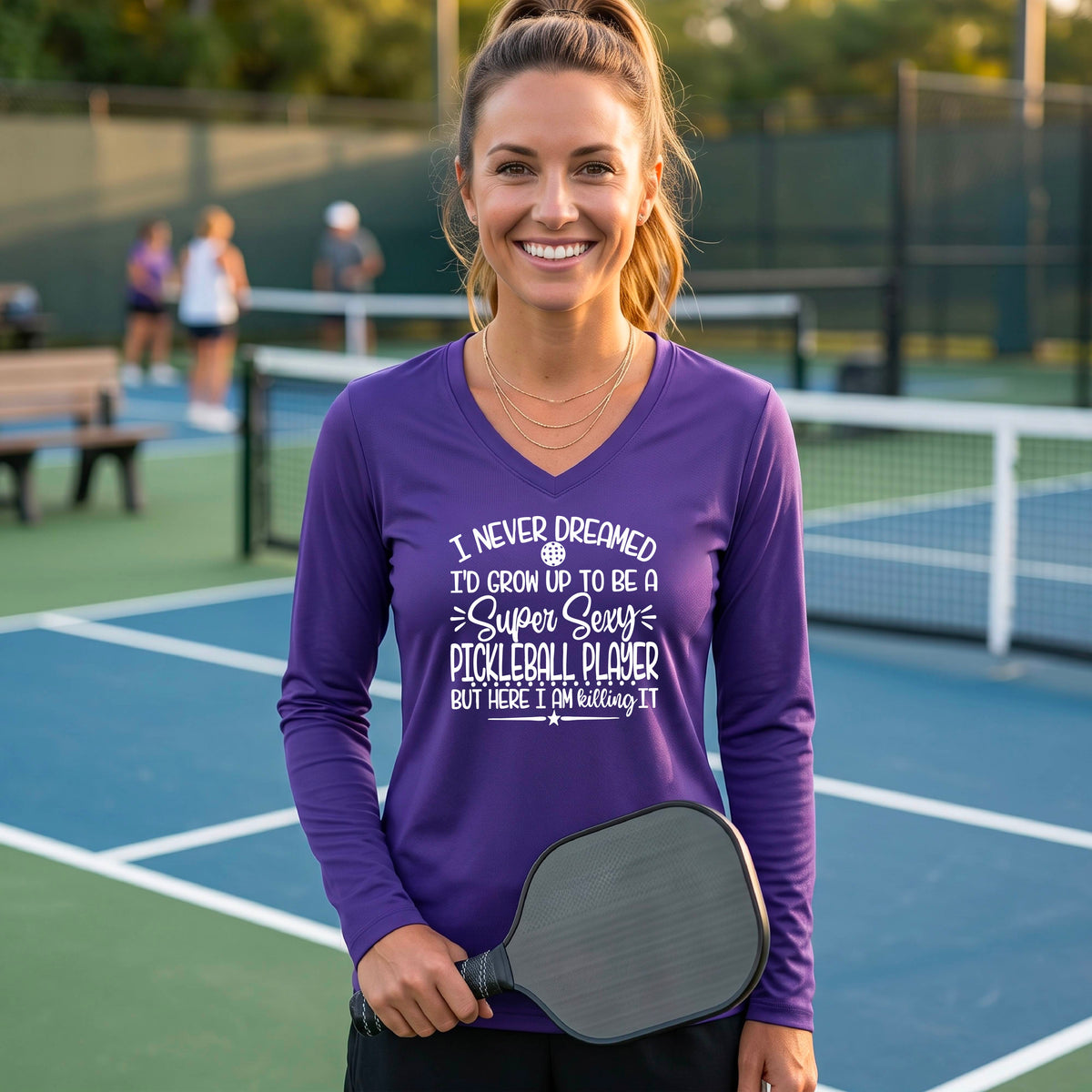 A woman on a pickleball court wearing a purple long-sleeve t-shirt with a white graphic of the text:  ‘I Never Dreamed I’d Grow Up To Be A Super Sexy Pickleball Player But Here I Am Killing It!’, standing on a pickleball court.