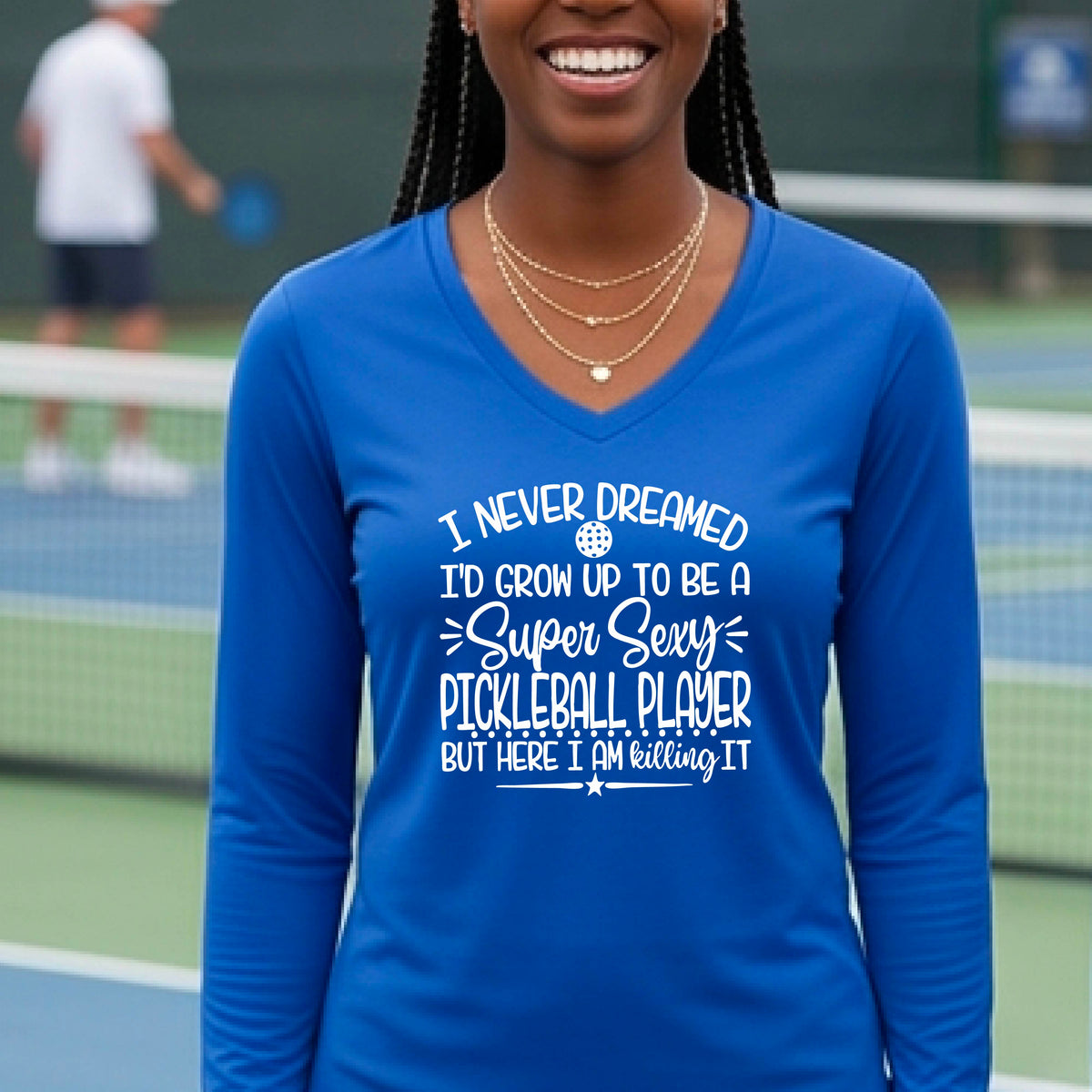 A woman on a pickleball court wearing a royal blue long-sleeve t-shirt with a white graphic of the text:  ‘I Never Dreamed I’d Grow Up To Be A Super Sexy Pickleball Player But Here I Am Killing It!’, standing on a pickleball court.