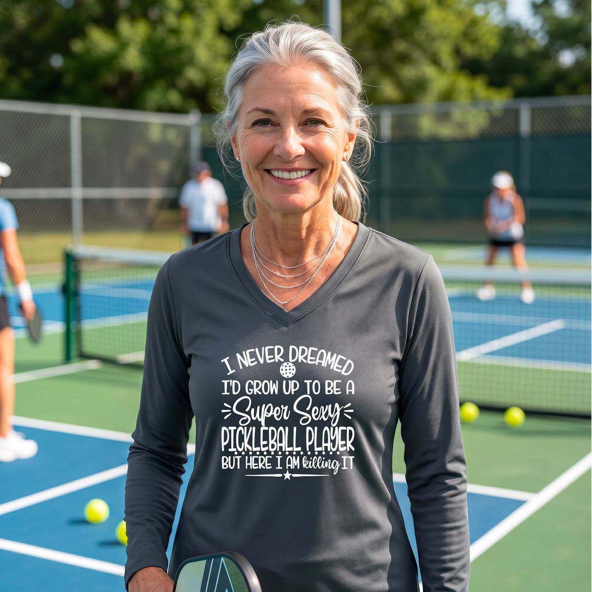A woman on a pickleball court wearing a dark gray long-sleeve t-shirt with a white graphic of the text:  ‘I Never Dreamed I’d Grow Up To Be A Super Sexy Pickleball Player But Here I Am Killing It!’, standing on a pickleball court.