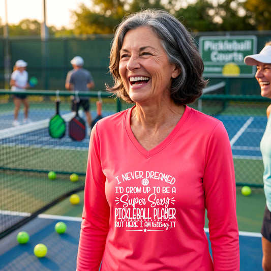 A woman on a pickleball court wearing a bright coral pink long-sleeve t-shirt with a white graphic of the text:  ‘I Never Dreamed I’d Grow Up To Be A Super Sexy Pickleball Player But Here I Am Killing It!’, standing on a pickleball court.