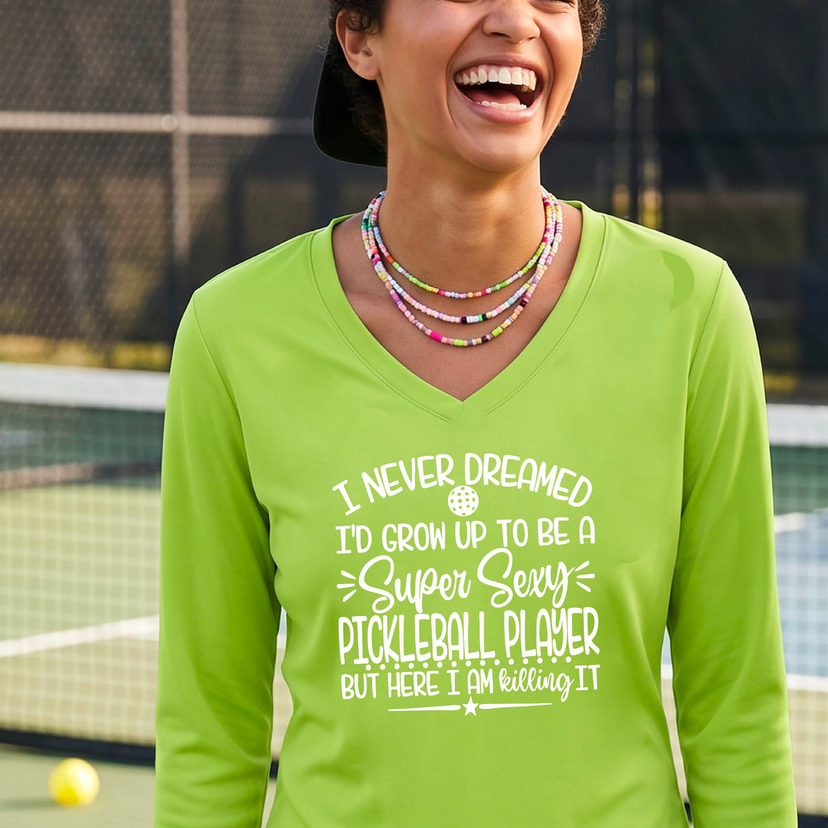 A woman on a pickleball court wearing a lime green long-sleeve t-shirt with a white graphic of the text:  ‘I Never Dreamed I’d Grow Up To Be A Super Sexy Pickleball Player But Here I Am Killing It!’, standing on a pickleball court.