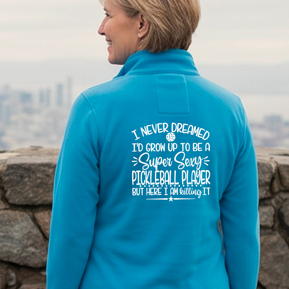 A woman on a pickleball court facing backwards wearing a bright blue quarter-zip tshirt with a white graphic of the text:  ‘I Never Dreamed I’d Grow Up To Be A Super Sexy Pickleball Player But Here I Am Killing It!’, standing on a pickleball court.