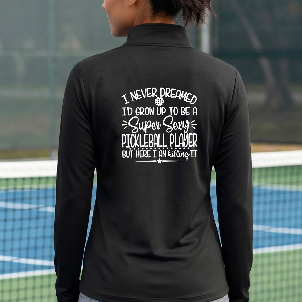A woman on a pickleball court facing backwards wearing a black quarter-zip tshirt with a white graphic of the text:  ‘I Never Dreamed I’d Grow Up To Be A Super Sexy Pickleball Player But Here I Am Killing It!’, standing on a pickleball court.