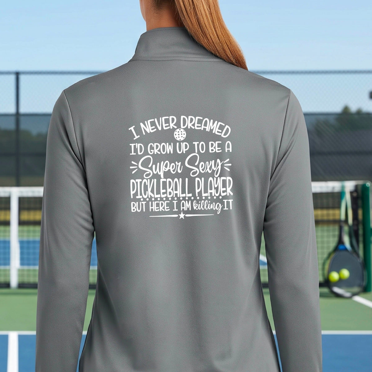 A woman on a pickleball court facing backwards wearing a gray quarter-zip tshirt with a white graphic of the text:  ‘I Never Dreamed I’d Grow Up To Be A Super Sexy Pickleball Player But Here I Am Killing It!’, standing on a pickleball court.