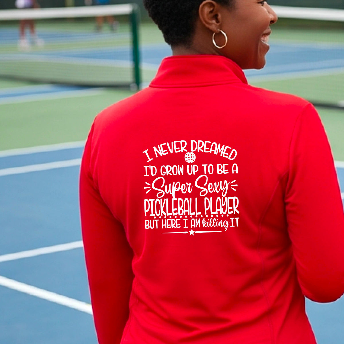 A woman on a pickleball court facing backwards wearing a red quarter-zip tshirt with a white graphic of the text:  ‘I Never Dreamed I’d Grow Up To Be A Super Sexy Pickleball Player But Here I Am Killing It!’, standing on a pickleball court.