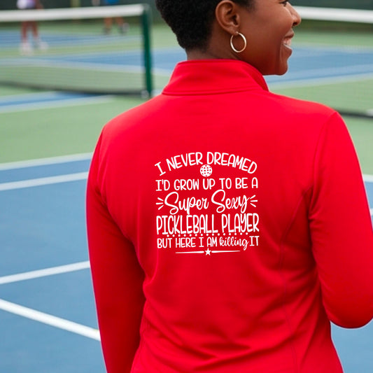 A woman on a pickleball court facing backwards wearing a red quarter-zip tshirt with a white graphic of the text:  ‘I Never Dreamed I’d Grow Up To Be A Super Sexy Pickleball Player But Here I Am Killing It!’, standing on a pickleball court.