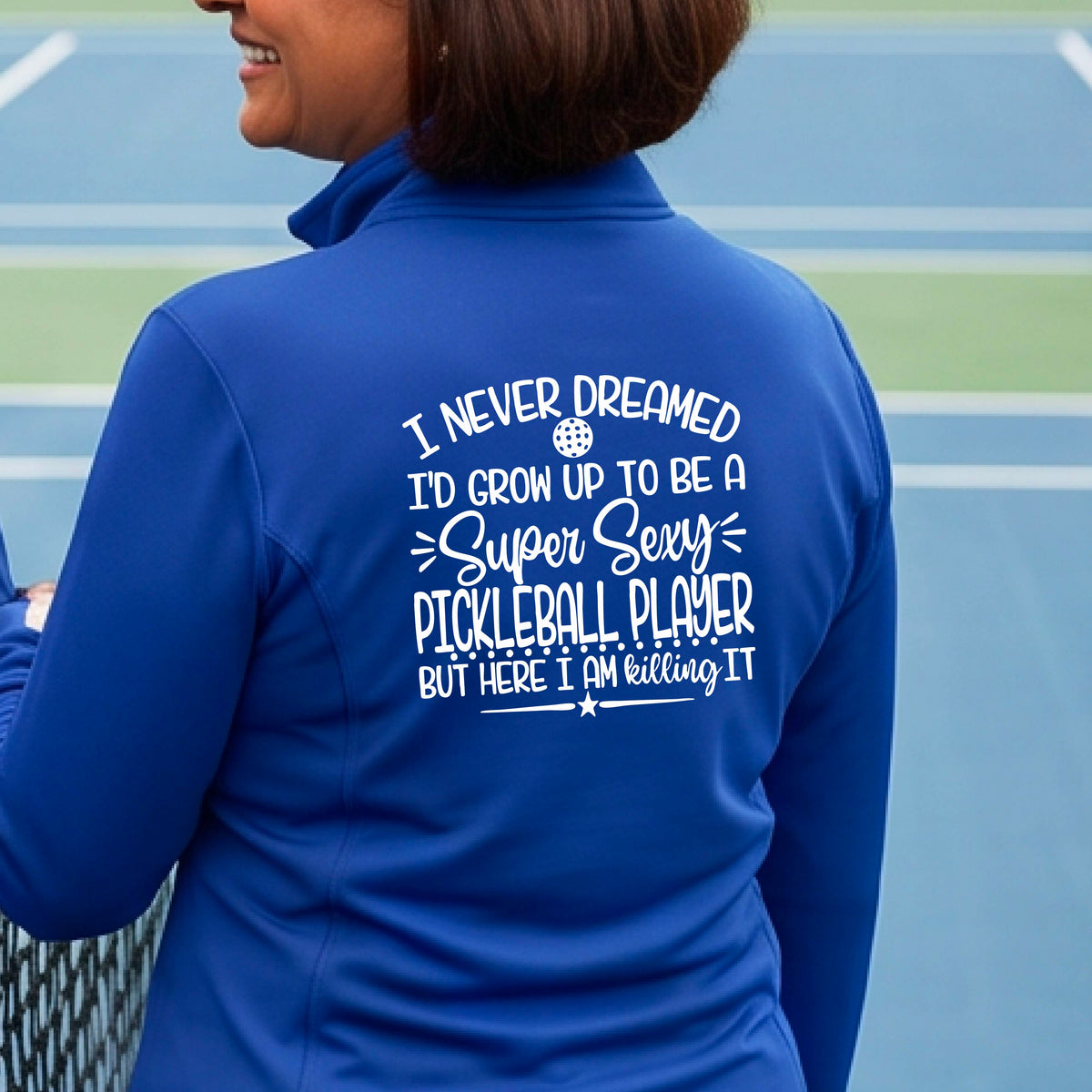 A woman on a pickleball court facing backwards wearing a royal blue quarter-zip tshirt with a white graphic of the text:  ‘I Never Dreamed I’d Grow Up To Be A Super Sexy Pickleball Player But Here I Am Killing It!’, standing on a pickleball court.