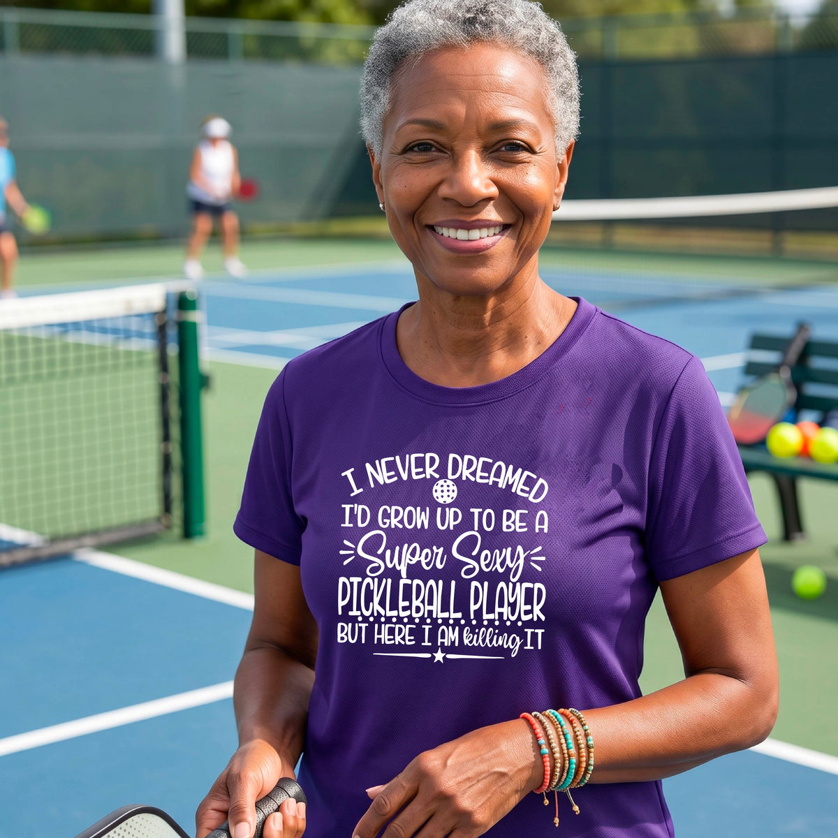 A woman on a pickleball court wearing a purple short-sleeve t-shirt with a white graphic of the text:  ‘I Never Dreamed I’d Grow Up To Be A Super Sexy Pickleball Player But Here I Am Killing It!’, standing on a pickleball court.