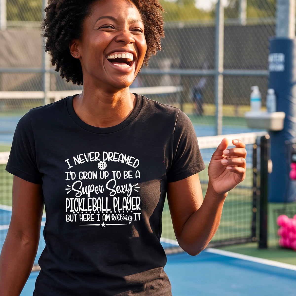 A woman on a pickleball court wearing a black short-sleeve t-shirt with a white graphic of the text:  ‘I Never Dreamed I’d Grow Up To Be A Super Sexy Pickleball Player But Here I Am Killing It!’, standing on a pickleball court.